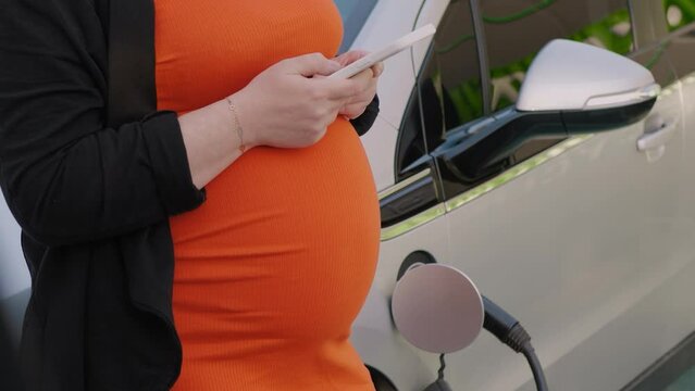 Unrecognizable pregnant woman in orange dress using the internet on her smartphone while waiting charging the battery at the electric car