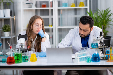 Young White Sciencetist woman and man sitting at desk with laptop and colorful liquid in experiment...
