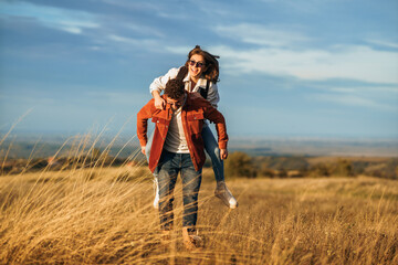 A boy is holding a girl on his back while walking through golden yellow meadow on a sunny day. They...