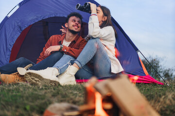 A couple is camping in nature, a girl is using binoculars to see something in the distance.