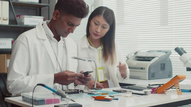 Medium shot of two technicians in white coats sitting at table and discussing broken smartphone at service centerMedium shot of two technicians in white coats sitting at table and discussing broken sm