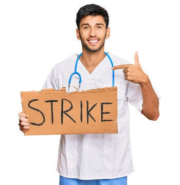 Young handsome man wearing doctor uniform holding strike banner cardboard pointing finger to one self smiling happy and proud
