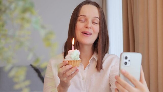 Cheerful brown haired woman with cake celebrating birthday on video call singing happy birthday song congratulating her friend with her holiday enjoying festive mood