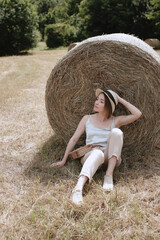 Naklejka na ściany i meble A beautiful girl is resting in a bale of hay on a summer morning field. Portrait of a young fashionable woman relaxing in haystacks, summer vacation in the countryside. . High quality photo