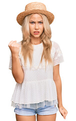 Young blonde girl wearing summer hat angry and mad raising fist frustrated and furious while shouting with anger. rage and aggressive concept.