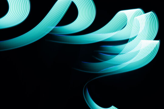 Turquoise green glow neon wave line of light as curls or swirl with smooth stripes on black background, pattern. Abstract background with flowing line in motion, light painting in vapor wave style.