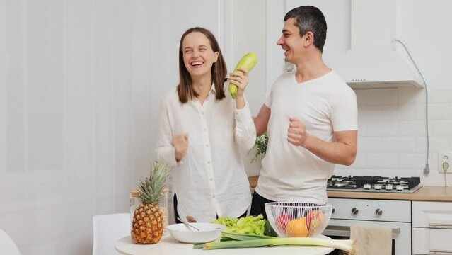 Funny cheerful couple dancing in kitchen and singing with zucchini as a microphone laughing happily having fin while preparing salad for dinner at home