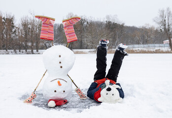 funny snowman stands upside down on frozen snowy lake. boy in polar bear hat lies next to him cheerfully raising legs.