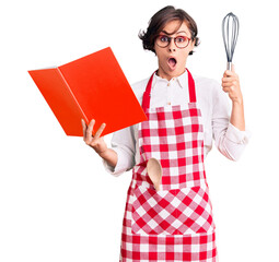 Beautiful young woman with short hair wearing professional baker apron reading cooking recipe book...