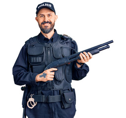 Young handsome man wearing police uniform holding shotgun looking positive and happy standing and smiling with a confident smile showing teeth