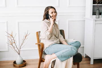 Millennial Woman Smiling, Speaking on Phone, Sitting on Chair, Looking Out Window at Free Space....