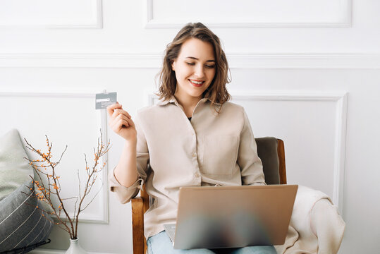 Joyful Millennial Woman Typing on Laptop, Seamlessly Using Credit Card for Online Shopping, Creating a Modern Image of Convenience and Enjoyment in Digital Retail Experience at Home.
