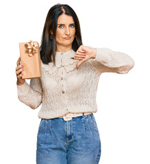 Middle age brunette woman holding gift with angry face, negative sign showing dislike with thumbs...