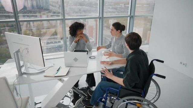 Paralyzed man works with colleagues. Guy in wheelchair has spine trauma discussing working project in office on meeting with multiethnic team women colleagues. Teamwork brainstorming, special needs.