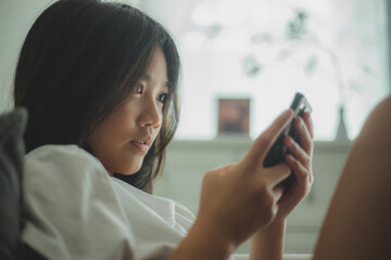 Happy Asian girl relaxing on a comfortable couch at home texting and messaging on a smartphone,...