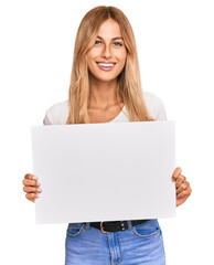 Fototapeta premium Beautiful blonde young woman holding blank empty banner looking positive and happy standing and smiling with a confident smile showing teeth