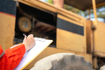 Action of an engineer hand is writing to perform inspection of the road roller or road compactor machinery engine part (as blurred background). Industrial working action scene, selective focus.  - obrazy, fototapety, plakaty