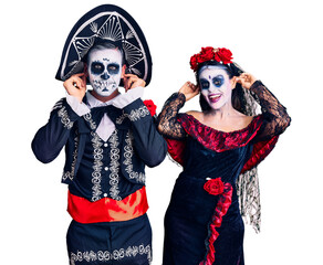 Young couple wearing mexican day of the dead costume over background smiling pulling ears with fingers, funny gesture. audition problem