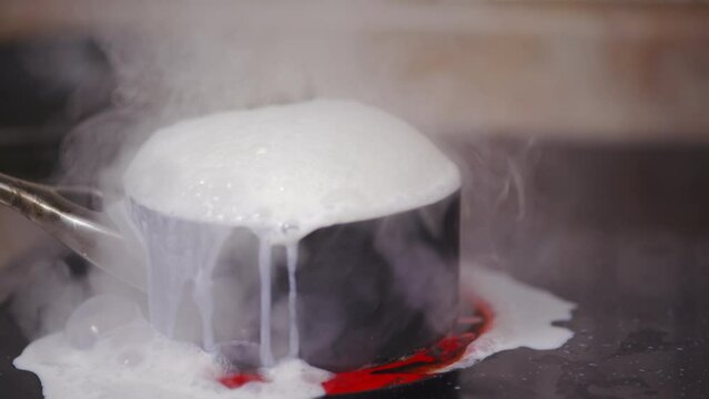 A pot of milk on a stovetop boils over, while steam escapes and milk bubbles and sticks to the top of the oven.