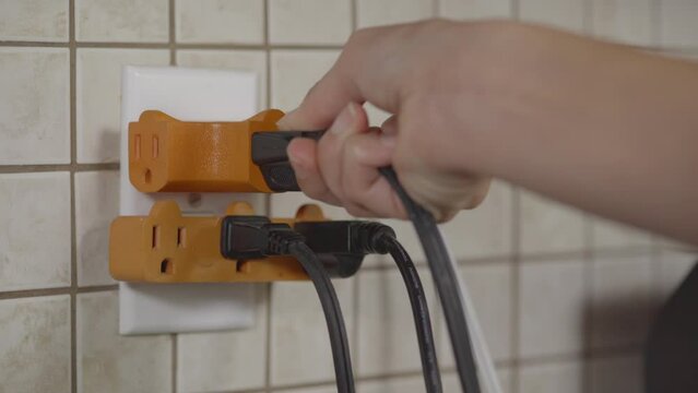 An outlet is fitted with plug splutters and several cords are plugged in already. A hand plugs in another cord to the outlet.