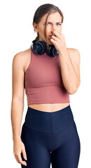 Beautiful caucasian young woman wearing gym clothes and using headphones smelling something stinky and disgusting, intolerable smell, holding breath with fingers on nose. bad smell - obrazy, fototapety, plakaty