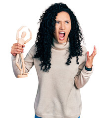 Young hispanic woman with curly hair holding small wooden manikin crazy and mad shouting and yelling with aggressive expression and arms raised. frustration concept.