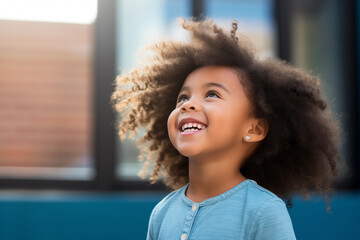 Portrait of a little girl in blue shirt looking up thinking, isolated on blue studio background. Close up young african american girl smiling and looking up