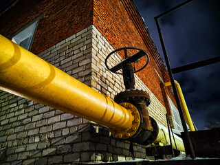 A large yellow pipe with a huge valve near the brick wall of the boiler room at night or in the...