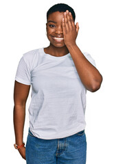 Young african american woman wearing casual white t shirt covering one eye with hand, confident smile on face and surprise emotion.