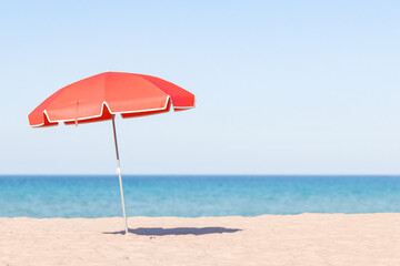 single red beach umbrella standing on a sandy beach with a clear blue sky and calm ocean in the background, peaceful beach day concept.