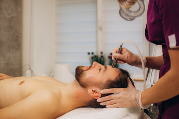 The cosmetologist makes the procedure Microdermabrasion of the facial skin of a man in beauty salon.