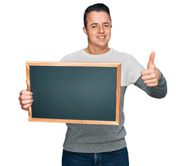 Handsome young man holding blackboard smiling happy and positive, thumb up doing excellent and approval sign