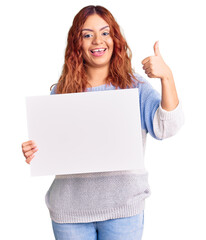 Young latin woman holding blank empty banner smiling happy and positive, thumb up doing excellent and approval sign