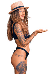 Young hispanic woman with tattoo wearing bikini and summer hat pointing aside with hands open palms showing copy space, presenting advertisement smiling excited happy