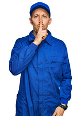 Bald man with beard wearing builder jumpsuit uniform asking to be quiet with finger on lips....