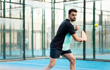 A grown man in sportswear is playing paddle tennis on a blue outdoor court. The boy is about to hit the ball with his racquet. Concept of men playing padel. Play padel outdoors.