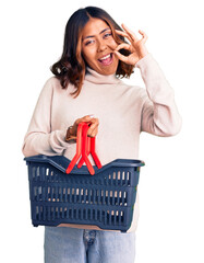 Young beautiful mixed race woman holding supermarket shopping basket doing ok sign with fingers, smiling friendly gesturing excellent symbol