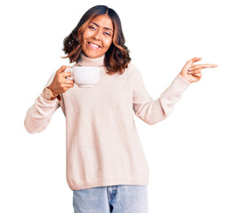 Young beautiful mixed race woman holding a cup of coffee smiling happy pointing with hand and...