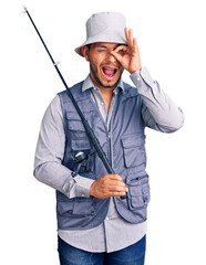 Handsome latin american young man wearing fisherman equipment smiling happy doing ok sign with hand on eye looking through fingers