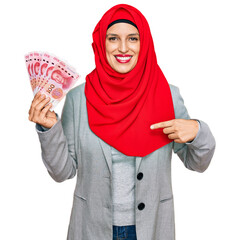 Beautiful hispanic woman wearing islamic hijab holding chinese yuan banknotes pointing finger to one self smiling happy and proud