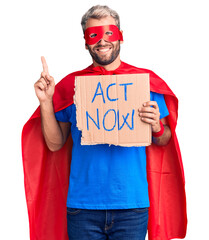 Young blond man wearing super hero costume holding act now cardboard banner surprised with an idea...