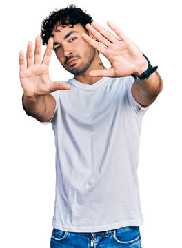 Hispanic young man with beard wearing casual white t shirt doing frame using hands palms and fingers, camera perspective