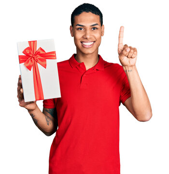 Young hispanic man holding gift smiling with an idea or question pointing finger with happy face, number one