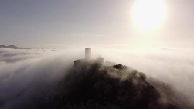 captivating drone zooms in on the majestic ruins of a castle atop a mountain. Mist weaves through ancient stones as the backlit sun creates a historic and mystical atmosphere in this compelling aerial