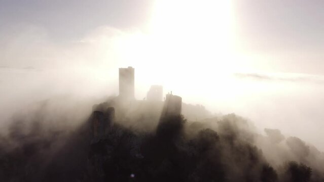 captivating drone zooms in on the majestic ruins of a castle atop a mountain. Mist weaves through ancient stones as the backlit sun creates a historic and mystical atmosphere in this compelling aerial