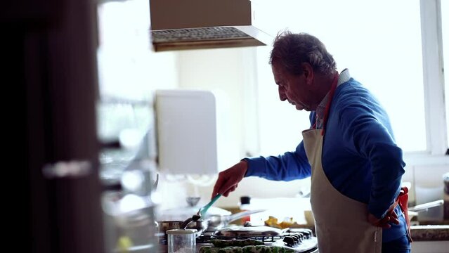 Senior chef cooking food by kitchen stove, candid authentic elderly retired man preparing meal for family wearing apron and blue sweater concentrated and cultivating hobby