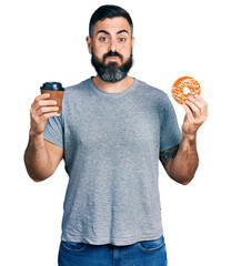 Hispanic man with beard eating doughnut and drinking coffee puffing cheeks with funny face. mouth...