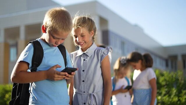 Children stand in schoolyard and look at smartphone.students with backpacks at school.children play smartphones in school yard after school.students listen to music on smartphone.happy family concept