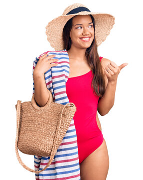 Young beautiful latin girl wearing swimwear and summer hat holding beach towel and bag pointing thumb up to the side smiling happy with open mouth