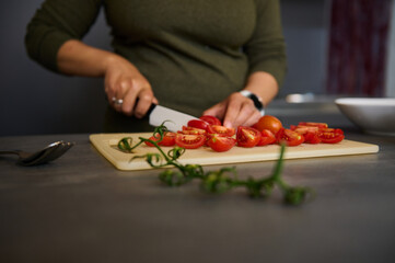 Obraz na płótnie Canvas Close-up hands of female chef using kitchen knife, chopping tomatoes on wooden cutting board, standing at kitchen table
